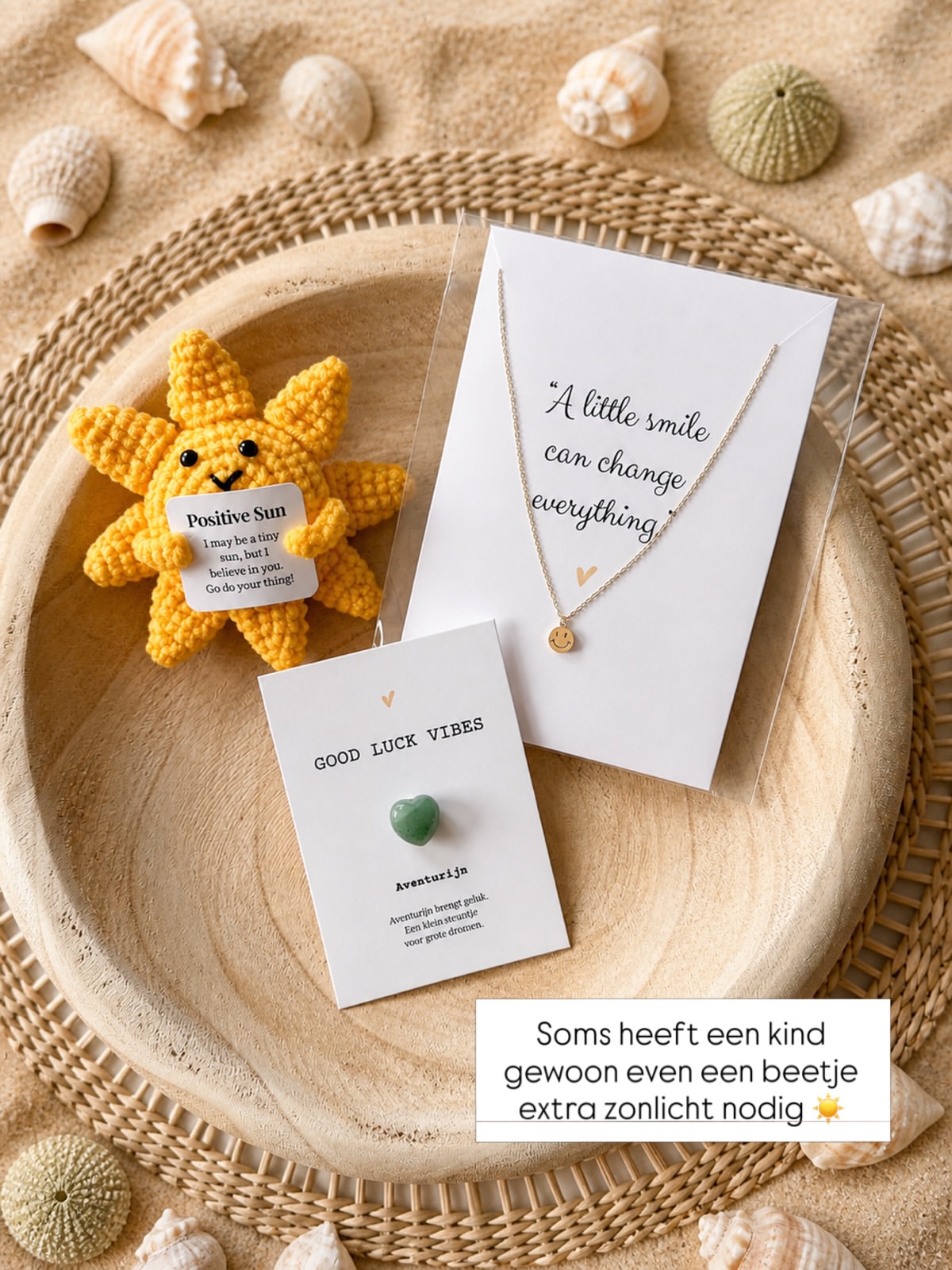 Yellow crochet sun plush with a 'Positive Sun' tag beside a necklace on a white card with 'A little smile can change everything' text, on a wicker mat.