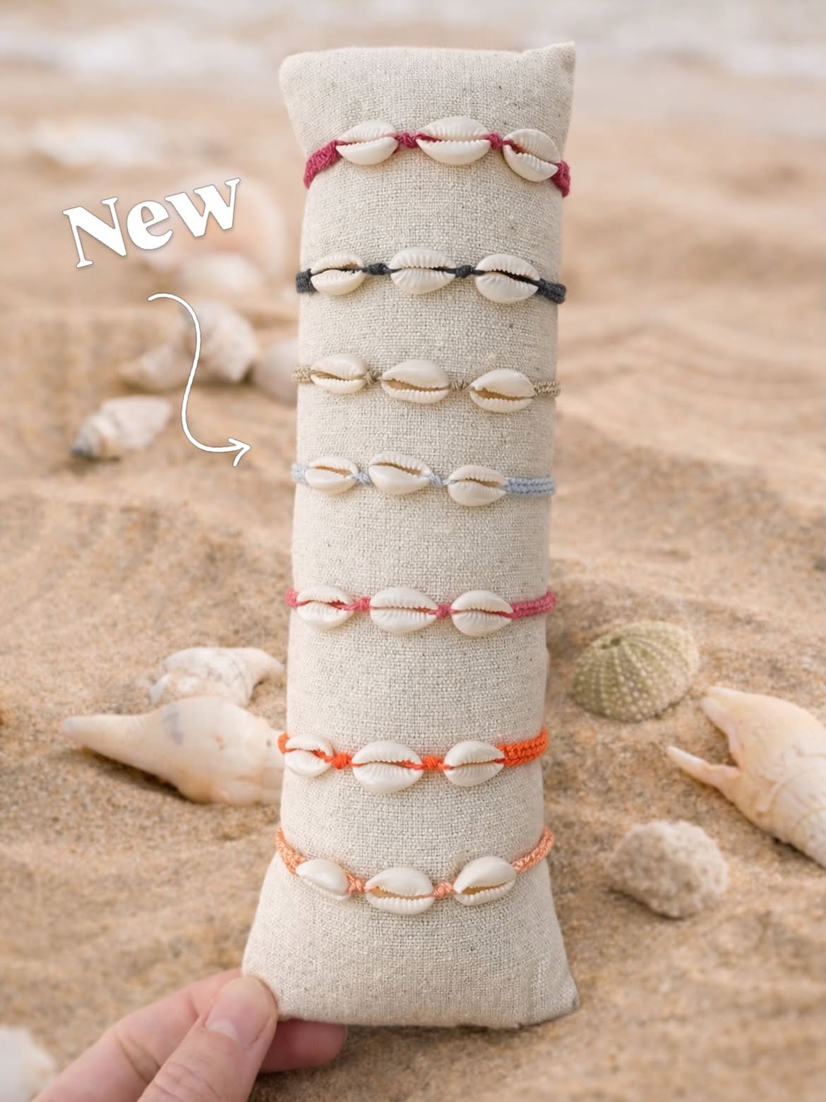 Beige fabric cone wrapped with multiple shell bracelets in red, black, gray, and orange on a sandy beach; a 'New' label is drawn on the left.