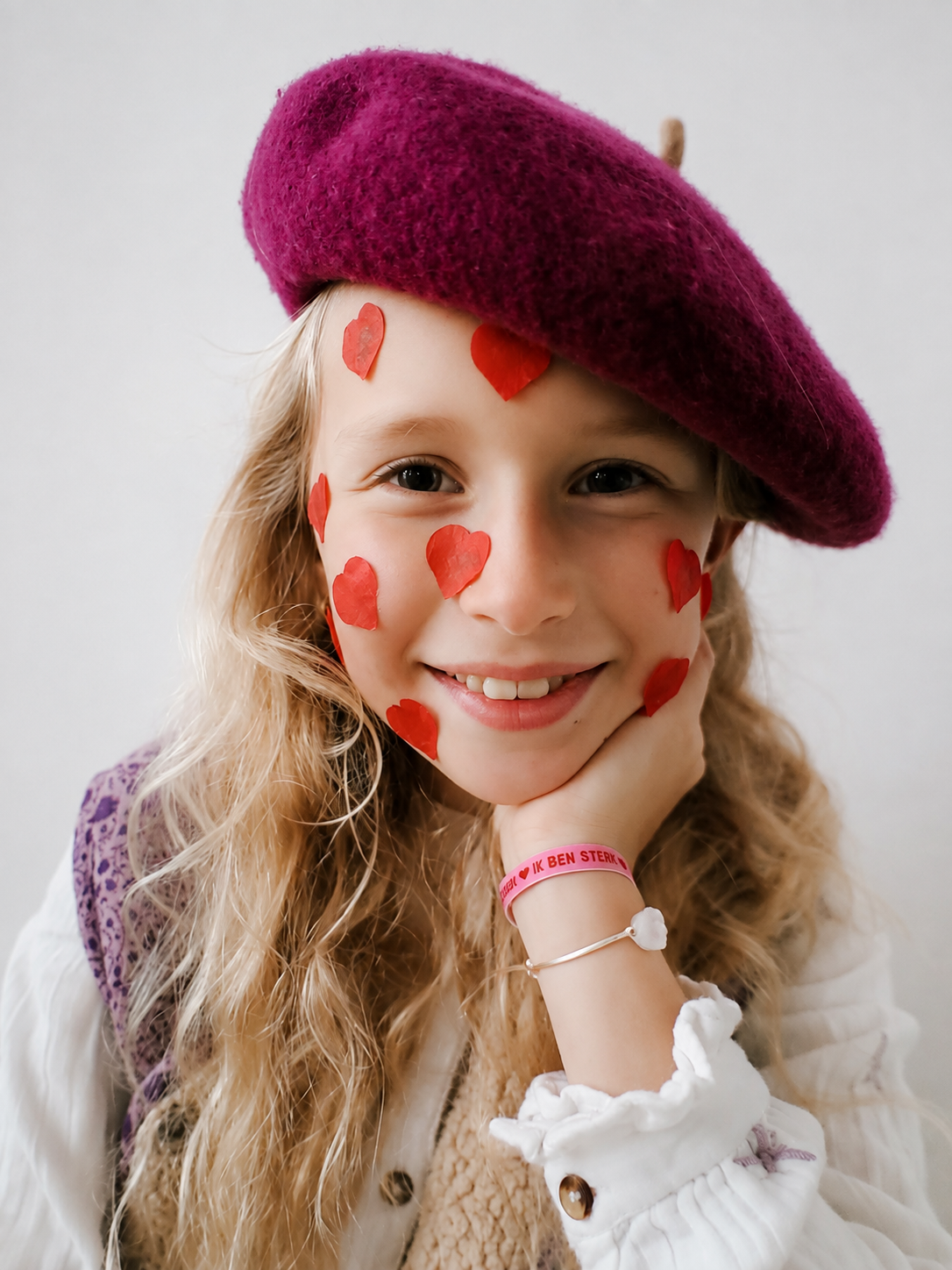 Smiling girl with long wavy hair wearing a purple beret and red heart stickers on her cheeks and forehead.
