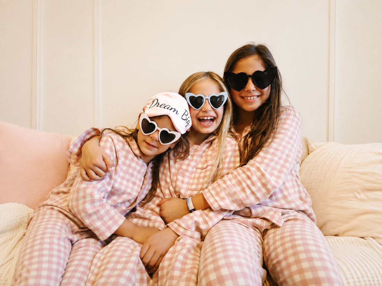 Three girls in matching pink gingham pajamas sit close together on a beige sofa, wearing heart-shaped sunglasses and smiling.